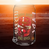 View of SCANDINORDIC HAPPY HOUR can glass from front, featuring a skull shaped like a can at the top, with colours red and white overlaid in similar style to the Danish flag. Above the skull the words in Viking Font, "HAPPY HOUR" and below, "VIKE STYLE". The glass is depicted over a background of the sea at sunset. Other sides of the glass design motifs are partially visible.